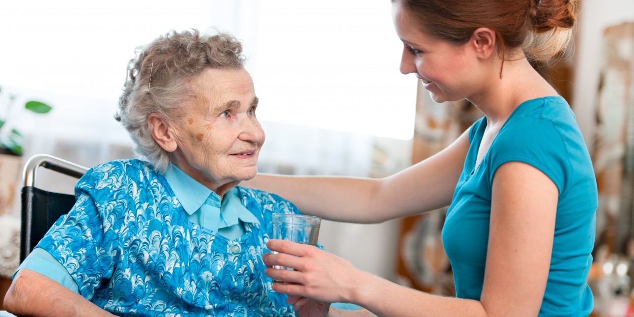 young woman helping elderly woman sit up, both individuals are smiling